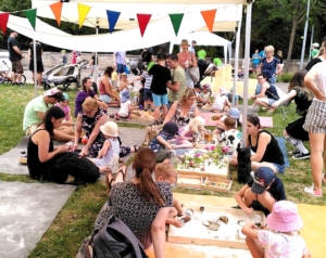 Viele Familien mit kleinen Kindern sitzen an flachen Holzkästen, in denen Spielmatrial liegt unter einem Pavillion im Freien